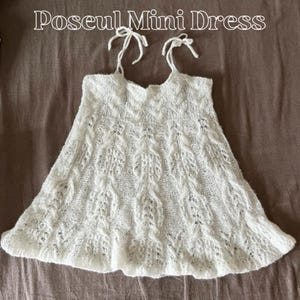 Puede incluir: Un minivestido blanco de punto con falda acampanada y delicadas tiras atadas. El vestido presenta un patrón texturizado de tejido abierto y el texto "Poseul Mini Dress" en la parte superior.