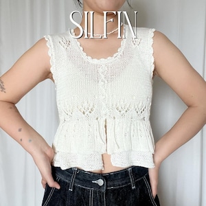 Può includere: Top senza maniche in maglia color crema con scollatura smerlata e orlo a balze. Il top presenta una parte anteriore con bottoni e dettagli traforati. La parola "SILFIN" è visualizzata nella parte superiore dell'immagine.