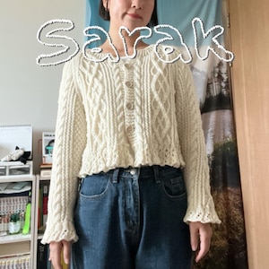 May include: A cream-coloured cable-knit cardigan with long sleeves and a button-up front. The cardigan has a ruffled hem and cuffs. The word "Sarak" is written in a cursive font across the top of the image. The person is wearing blue jeans.