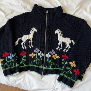 Könnte beinhalten: Dunkelblauer Strick-Cardigan mit Reißverschluss und hohem Kragen. Er zeigt zwei weiße Pferdesilhouetten und ein Blumenmuster. Der Cardigan hat einen silbernen Reißverschluss und hellblaue Bündchen.
