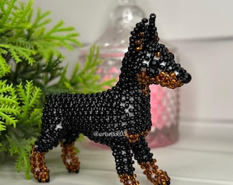 Può includere: Una piccola statuetta di cane tridimensionale realizzata con minuscole perline nere e marroni. La figura presenta orecchie a punta, un muso definito e quattro zampe. Il testo @ariwixx03 è visibile sul lato del corpo, integrato nella lavorazione.