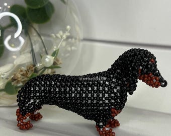 Può includere: Una statuetta di cane bassotto realizzata a mano con piccole perline nere, bianche e marrone rossiccio. Il cane presenta un corpo lungo, zampe corte e un muso dettagliato. Il testo @ariwix03 è visibile sul fianco del corpo.