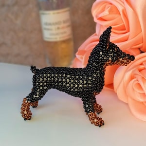 May include: A beaded miniature dog figurine in black, brown, and cream. The dog is posed in a standing position, with its legs and snout in brown. The background includes a blurred bottle and peach-coloured roses.