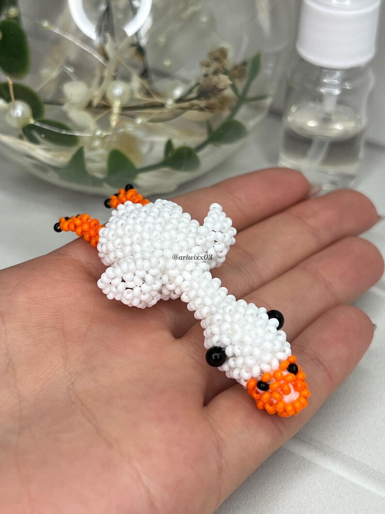 Pu&ograve; includere: Una piccola statuetta di perline fatta a mano a forma di oca, realizzata con minuscole perline bianche per il corpo e la testa, con perline arancioni vivaci per le zampe e il becco. L'oca presenta due piccole perline nere per gli occhi e il testo @ariwixx08.