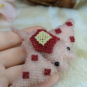 May include: A beaded stingray pin in shades of pink and red. The stingray is made of small pink beads, with a central red square and smaller red accents. Black beads form the eyes.