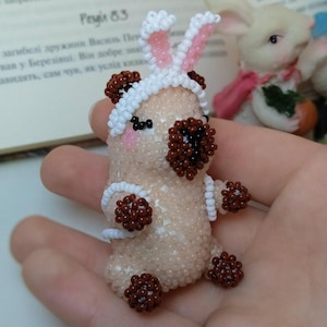 May include: A handmade beaded figurine of a bunny with brown and white accents. The bunny has a white headband with pink-lined ears and brown bead accents. The figurine is held in a hand.