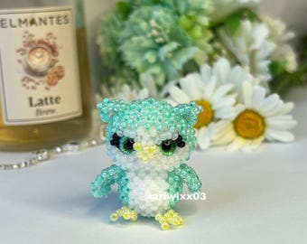 Può includere: Una piccola statuetta a forma di gufo realizzata con minuscole perline color verde menta e bianco. Il gufo presenta grandi occhi verdi con ciglia nere, un becco giallo e zampe gialle. Il testo @ariwixx03 è visibile sulla parte inferiore.