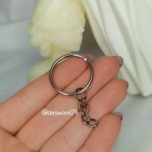 Pu&ograve; includere: Un portachiavi in metallo color argento composto da un anello circolare diviso attaccato a una corta catena a tre maglie. Il testo @ariwixx03 &egrave; stampato in bianco sulla superficie dell'anello metallico. Il portachiavi ha una finitura lucida.