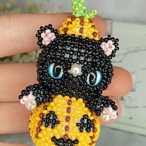 May include: A handmade beaded Halloween cat figurine. The black cat has blue eyes and is wearing a pumpkin hat. The cat is sitting on a yellow pumpkin. The figurine is made with small beads and has a detailed design.