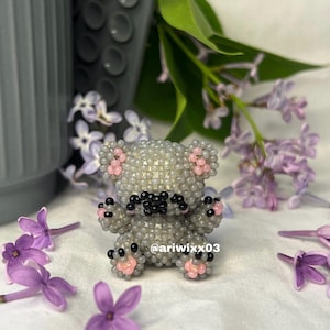 May include: A beaded grey koala bear figurine with pink and black accents. The koala has small black eyes and a black nose. The paws and ears are pink. The figurine is surrounded by purple flowers and green leaves.