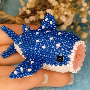 May include: A handmade beaded whale shark figurine. The blue body is detailed with white spots, and the mouth is pink. The eyes are black. The figurine is held against a light blue background.