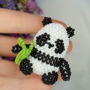 Puede incluir: Figura de oso panda hecha a mano con cuentas blancas y negras, sosteniendo un tallo de bambú verde con cuentas. El panda tiene ojos, orejas y pies negros. La figura se sostiene en una mano.