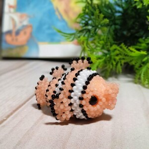 Puede incluir: Una figura de pez payaso hecha con cuentas. El pez tiene un cuerpo melocot&oacute;n con rayas blancas y negras y detalles de cuentas negras. El pez tiene un ojo grande y negro. La figura est&aacute; sobre una superficie de madera clara.