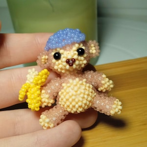 May include: A small beaded monkey wearing a blue hat and holding a yellow banana. The monkey is made of light pink and yellow beads.