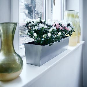 Könnte beinhalten: Ein weißes Fensterbrett mit einem silbernen Pflanztopf, gefüllt mit weißen und rosa Blüten. Zwei klare Glasvasen mit goldener Tönung stehen auf dem Fensterbrett.