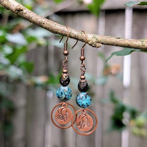 May include: Copper-toned drop earrings with a mountain and moon motif. The earrings feature blue and black beads and are suspended from a branch. The earrings have a rustic, handmade aesthetic.