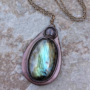 May include: A teardrop-shaped pendant necklace with a large, iridescent labradorite stone. The stone displays shades of blue, green, and gold. The pendant is framed in a copper-colored metal and hangs from a matching chain.