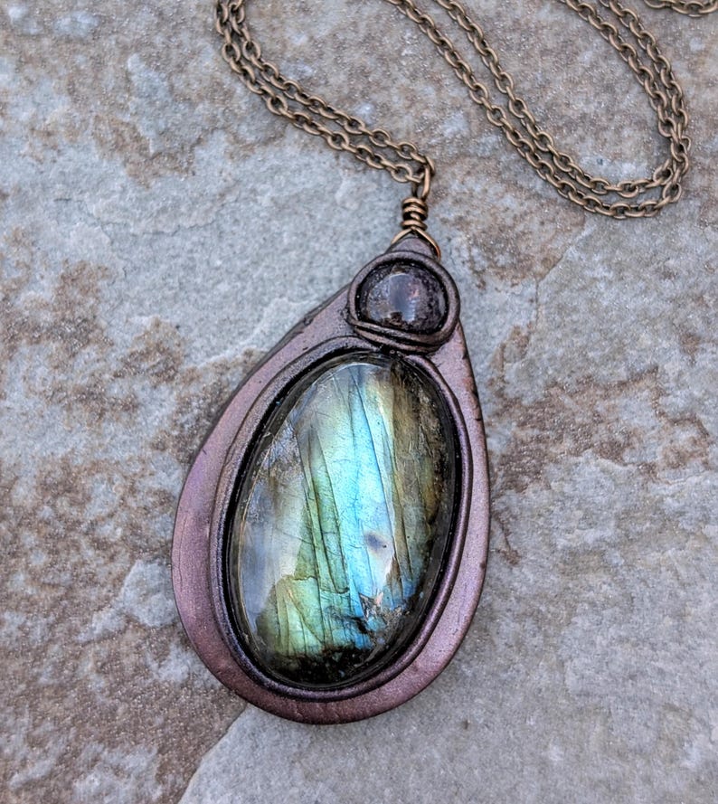 May include: A teardrop-shaped pendant necklace with a large, iridescent labradorite stone. The stone displays shades of blue, green, and gold. The pendant is framed in a copper-colored metal and hangs from a matching chain.
