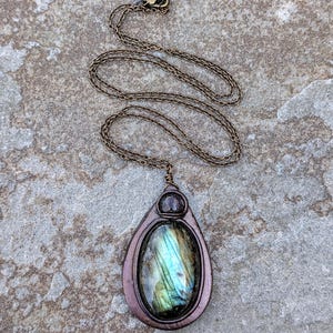 May include: A handcrafted pendant necklace featuring a teardrop-shaped pendant with a labradorite stone. The stone displays iridescent blue and green hues. The pendant is framed in a bronze-colored metal and hangs from a matching chain.