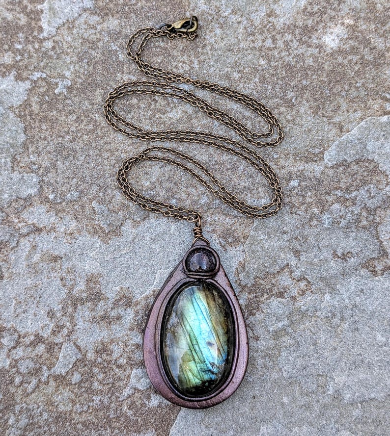 May include: A handcrafted pendant necklace featuring a teardrop-shaped pendant with a labradorite stone. The stone displays iridescent blue and green hues. The pendant is framed in a bronze-colored metal and hangs from a matching chain.