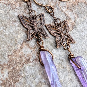 May include: A pair of dangle earrings featuring antique copper-toned butterfly charms connected to elongated, translucent light purple crystal points. The metal components have a dark, weathered finish, and the crystals are secured with wire wraps.