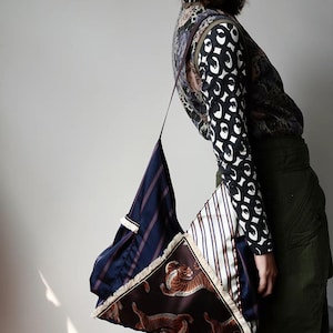 Puede incluir: Un bolso de hombro triangular único. El bolso es un patchwork de diferentes telas, incluyendo azul marino con rayas blancas, azul marino con rayas verticales y una tela marrón con ilustraciones de tigres. Los bordes están adornados con flecos.