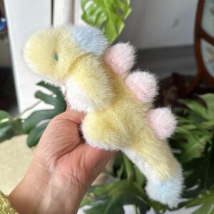 May include: A soft, plush toy in the shape of a whimsical creature. The toy features a yellow body with white accents, and light blue and pink details. The toy is held in a hand, with a green plant in the background.