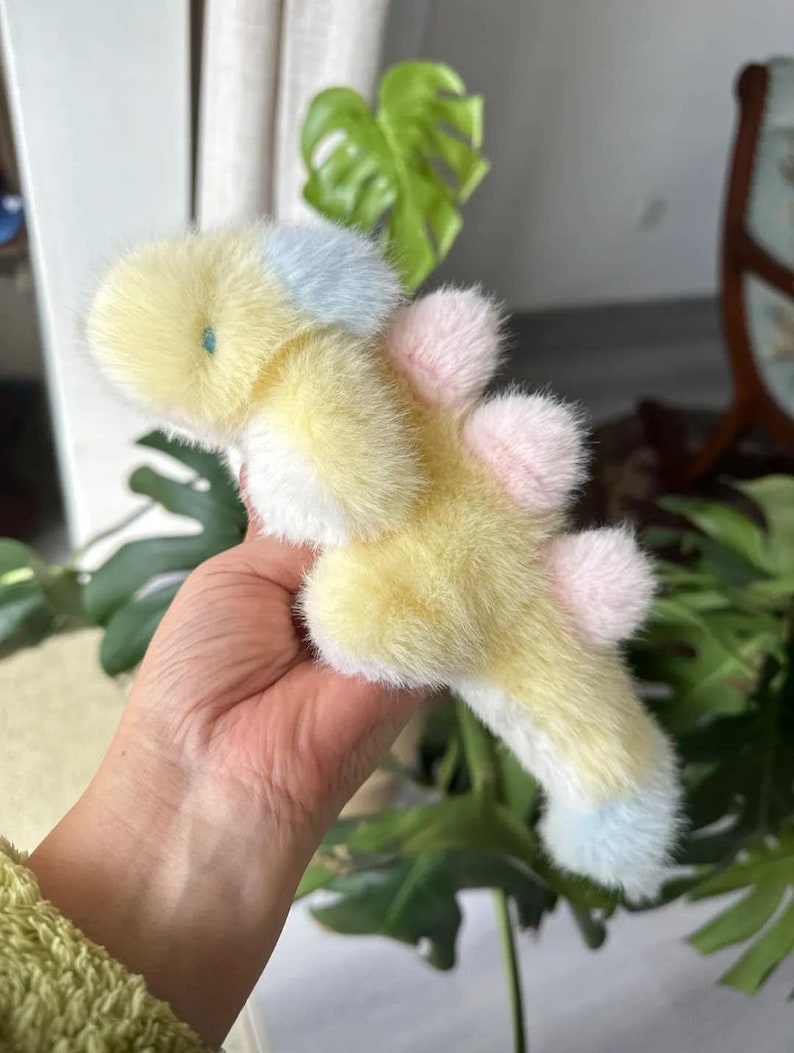May include: A soft, plush toy in the shape of a whimsical creature. The toy features a yellow body with white accents, and light blue and pink details. The toy is held in a hand, with a green plant in the background.