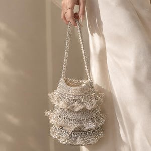 Op de afbeelding: Een metallic zilveren bucket bag met drie gelaagde ruches en een gevlochten handvat. De tas is gemaakt van een glinsterend, getextureerd gebreid materiaal dat het licht opvangt, wat zorgt voor een gelaagd, volumineus silhouet met een ronde bodem.