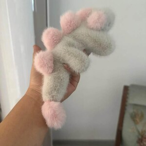 May include: A plush toy in the shape of a dinosaur, featuring a soft, fuzzy texture. The toy is primarily light gray with pink accents on the head, back, and tail. The dinosaur has a playful, whimsical design.