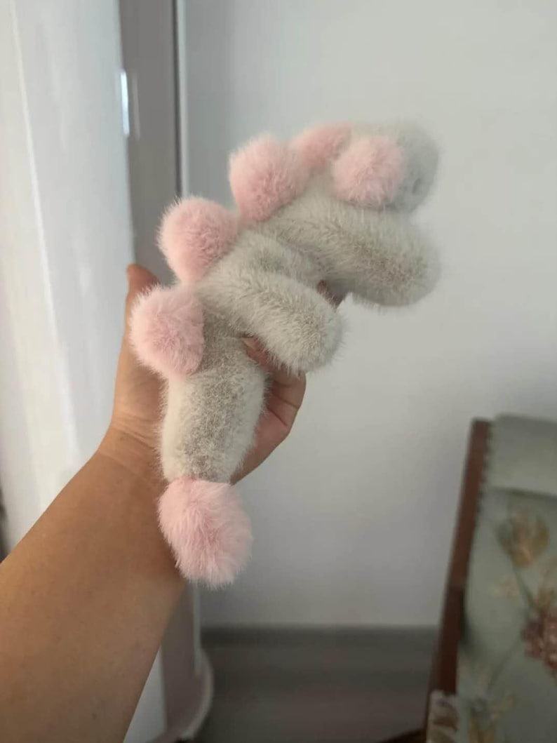 May include: A plush toy in the shape of a dinosaur, featuring a soft, fuzzy texture. The toy is primarily light gray with pink accents on the head, back, and tail. The dinosaur has a playful, whimsical design.