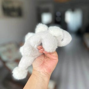 May include: A soft, white plush bunny toy with floppy ears and a small, round tail. The bunny is held in a hand, showcasing its fluffy texture and simple design. The toy is a classic children's item.
