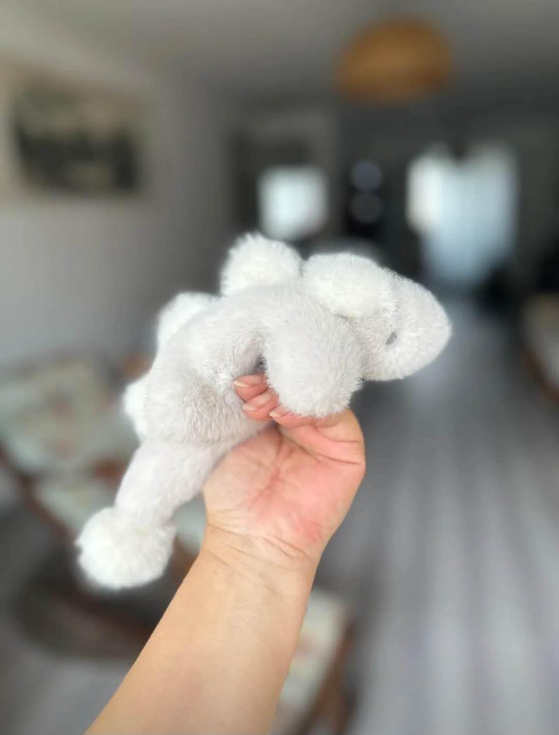May include: A soft, white plush bunny toy with floppy ears and a small, round tail. The bunny is held in a hand, showcasing its fluffy texture and simple design. The toy is a classic children's item.