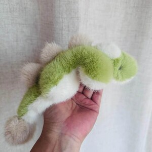 May include: A plush toy in the shape of a green and white dinosaur with a light brown tail and spikes. The toy is made of soft, fluffy material and is held in a person's hand.