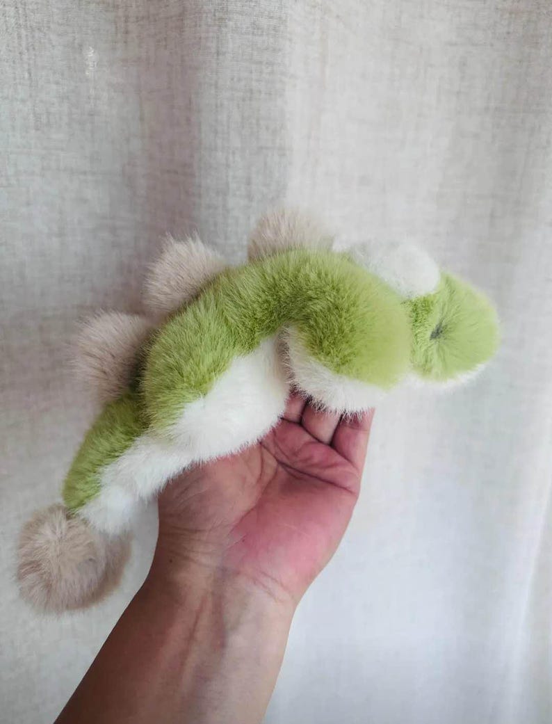 May include: A plush toy in the shape of a green and white dinosaur with a light brown tail and spikes. The toy is made of soft, fluffy material and is held in a person's hand.