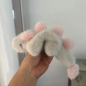 May include: A plush, light gray stuffed animal with pink accents. The toy has a fuzzy texture and features pink pom-poms and a pink face. The toy is held in a hand.