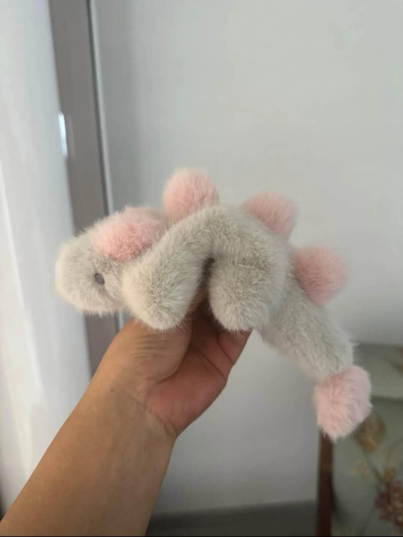 May include: A plush, light gray stuffed animal with pink accents. The toy has a fuzzy texture and features pink pom-poms and a pink face. The toy is held in a hand.