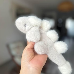 May include: A plush, light gray stuffed animal shaped like a creature with fluffy white accents. The toy has a soft, furry texture and a small, dark eye. The toy is held in a hand.