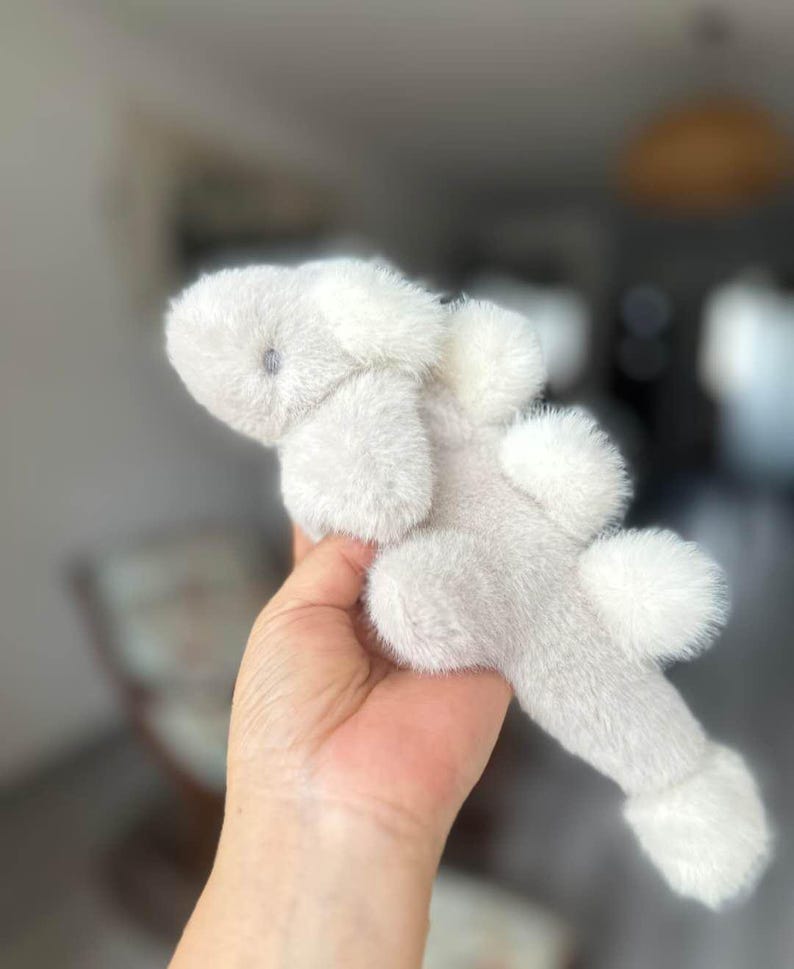 May include: A plush, light gray stuffed animal shaped like a creature with fluffy white accents. The toy has a soft, furry texture and a small, dark eye. The toy is held in a hand.