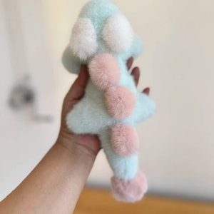 May include: A soft, plush toy in the shape of a star, with a light blue body and white and pink accents. The toy has a fluffy texture and is held in a person's hand.