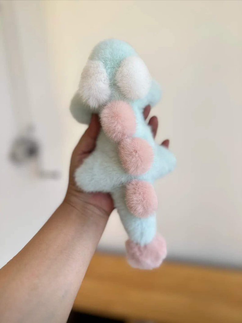 May include: A soft, plush toy in the shape of a star, with a light blue body and white and pink accents. The toy has a fluffy texture and is held in a person's hand.