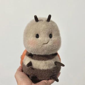 Puede incluir: Un juguete de peluche con forma de insecto con una cabeza redonda de color marrón claro, ojos negros y una pequeña sonrisa. El cuerpo está rayado con marrón y beige, con detalles en naranja. El juguete tiene pequeñas patas y antenas marrones oscuras.