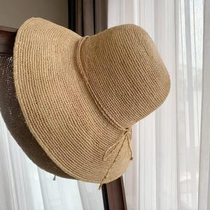 May include: A wide-brimmed straw hat in a natural tan colour, featuring a rounded crown and a woven texture. The hat includes a thin, matching straw cord tied in a small bow detail at the base of the crown, resting above the wide, flexible brim.