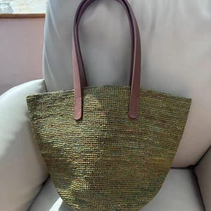 May include: A tote bag featuring a woven, textured body in variegated shades of olive green, brown, and yellow. The bag has two smooth, solid brown leather-like shoulder straps attached to the top edge with visible stitching at the base of each handle.