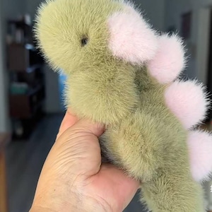 May include: A plush, green dinosaur toy with pink accents. The toy has a soft, furry texture and features pink pom-poms along its back and on its feet. The dinosaur is held in a hand, showcasing its size and design.