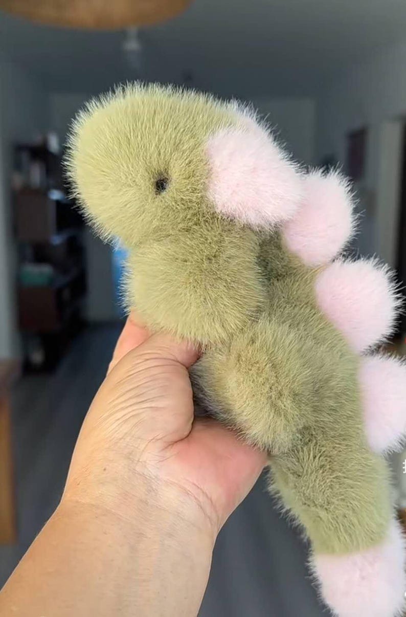 May include: A plush, green dinosaur toy with pink accents. The toy has a soft, furry texture and features pink pom-poms along its back and on its feet. The dinosaur is held in a hand, showcasing its size and design.