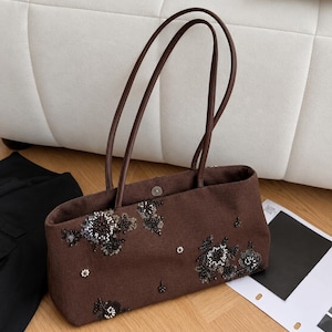 May include: A dark brown rectangular tote bag featuring two thin shoulder straps and a magnetic snap closure. The exterior is decorated with scattered floral-inspired embellishments made of black beads, silver sequins, and small white pearls.