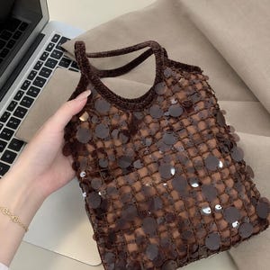 May include: A dark brown crochet handbag featuring a grid-like open weave pattern adorned with numerous circular, translucent brown sequins that catch the light. The bag has two integrated crochet handles and a structured, square-shaped body.