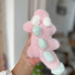 May include: A pink plush toy with white and light blue accents. The toy has a soft, furry texture and a unique shape, with white spots and light blue circular details. The toy is held in a hand, with a blurred background.