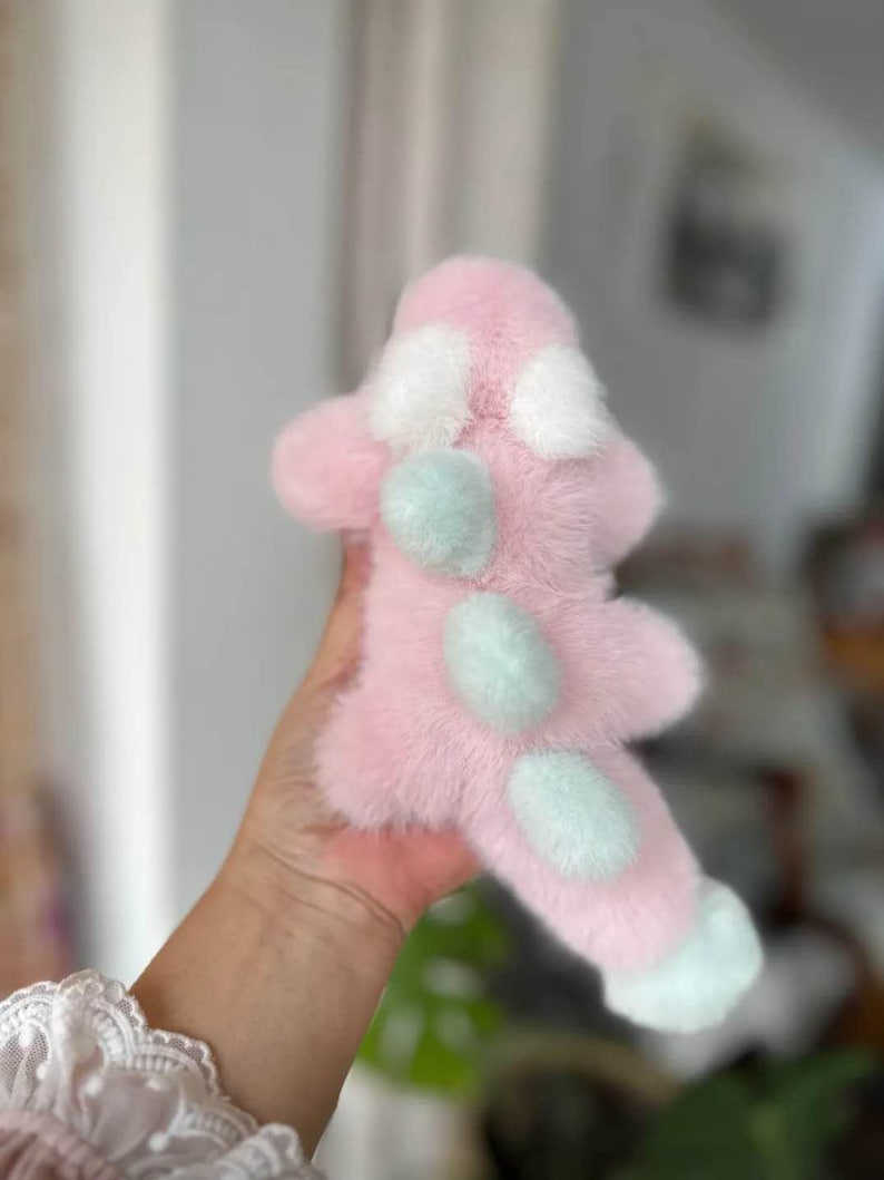 May include: A pink plush toy with white and light blue accents. The toy has a soft, furry texture and a unique shape, with white spots and light blue circular details. The toy is held in a hand, with a blurred background.